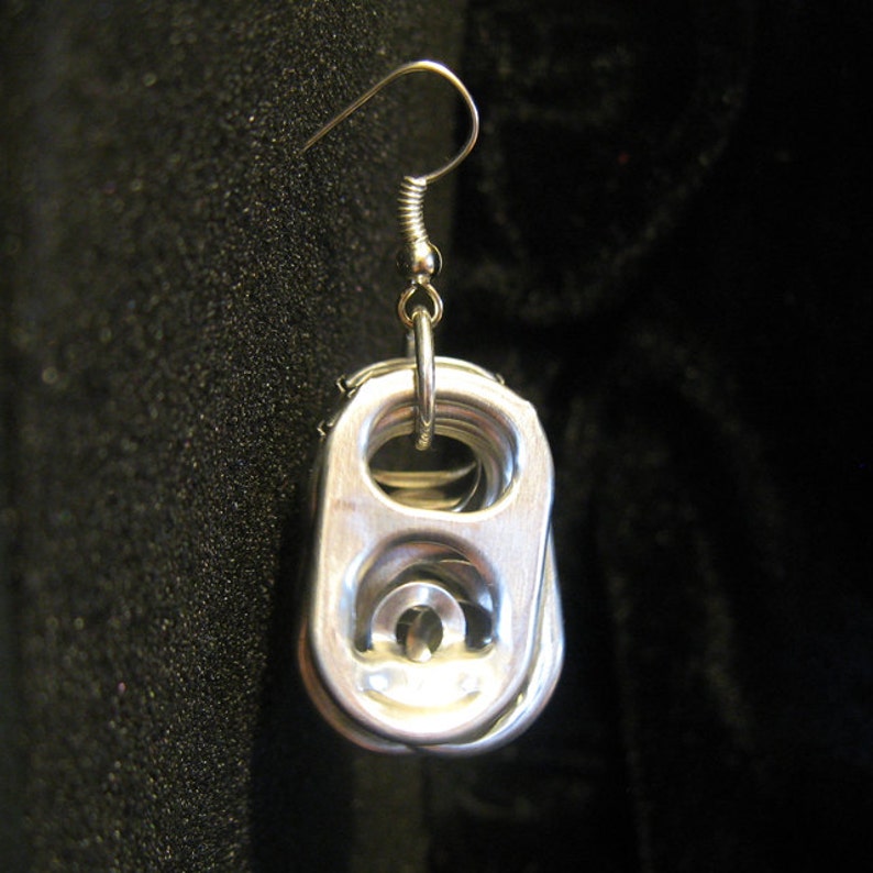 May include: A single silver-colored earring made from a recycled soda can tab. The earring features a hook and a circular ring connecting to the tab. The tab has a rounded rectangular shape with a raised center. The background is a dark, textured fabric.