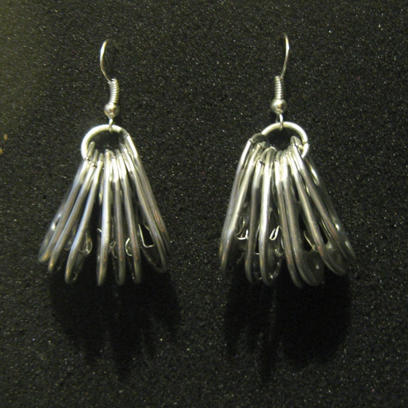 May include: A pair of silver-colored dangle earrings. Each earring features a cluster of silver metal pieces, arranged in a fan shape, suspended from a silver ear wire. The earrings are displayed against a dark background.