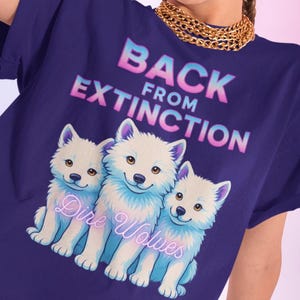 Op de afbeelding: Een paars T-shirt met een afbeelding van drie witte wolven en de tekst "Back from Extinction - Dire Wolves".