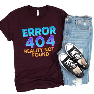 May include: A maroon t-shirt with the text "ERROR 404 REALITY NOT FOUND" in blue and yellow. The shirt is paired with light blue denim jeans and black canvas sneakers with white laces and soles. The items are arranged on a white background.