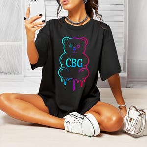 May include: Black t-shirt featuring a colorful outline of a gummy bear with the letters "CBG" in the center. The bear design has a gradient effect, transitioning from blue to pink. The shirt is a casual, oversized style.