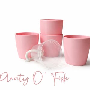 May include: A collection of pink plant pots, some stacked, with a clear plastic container and the text "Planty O' Fish" in a cursive font. The pots are cylindrical and have a smooth finish.