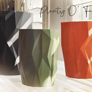 May include: Three geometric planters in black, olive green, and orange with a faceted surface texture. The text Planty O' Fish is visible in the upper right corner. These cylindrical containers feature a distinct angular design throughout.