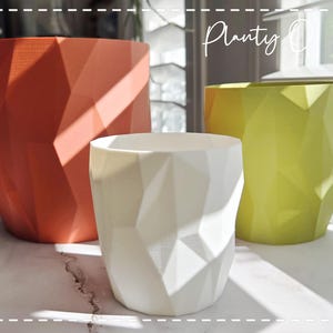 May include: Three geometric planters in orange, white, and lime green with a faceted, angular surface texture. The planters are arranged in a group, and the text "Planty O Fish" is visible in the upper right corner of the image.