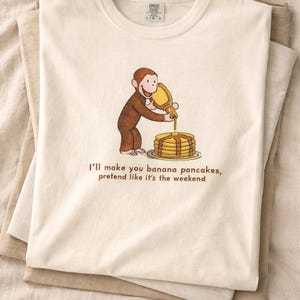 May include: Cream-colored t-shirt featuring a cartoon monkey pouring syrup onto a stack of pancakes. The text "I'll make you banana pancakes, pretend like it's the weekend" is printed below the illustration. The shirt is folded.