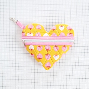 Może przedstawiać: Torebka z zamkiem błyskawicznym w kształcie serca, z żółtym materiałem zewnętrznym i różową podszewką. Materiał ma wzór białych truskawek na żółtym tle. Torebka ma srebrny zamek błyskawiczny i srebrną pętelkę do przymocowania breloka.