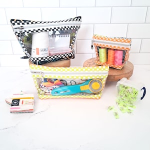 Pode incluir: Três bolsas de vinil transparente com fechos de correr e acabamento em tecido xadrez preto, branco, laranja e amarelo. As bolsas contêm artigos de costura, incluindo carretéis de linha, um cortador rotativo e clipes verde néon.