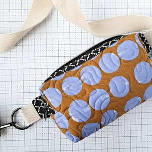 May include: A brown and blue quilted fabric bag with a white strap and a zipper closure. The bag has a pattern of blue circles on a brown background.