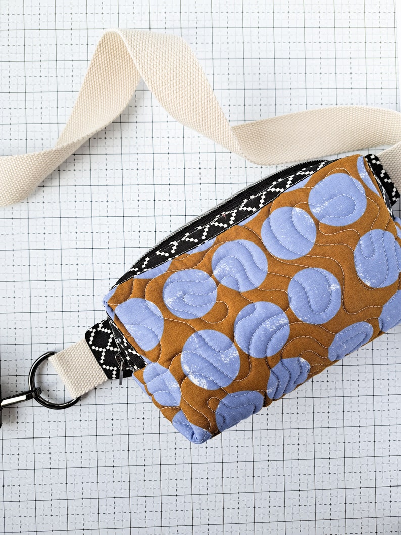 May include: A brown and blue quilted fabric bag with a white strap and a zipper closure. The bag has a pattern of blue circles on a brown background.