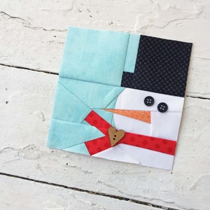 May include: A fabric snowman with a light blue top, black hat, and a red dotted scarf. The snowman has a carrot nose, black button eyes, and a wooden heart-shaped button. The design is on a white background.