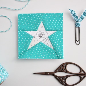 May include: A teal fabric square with a white star in the center. The star is made of smaller fabric squares with different patterns. The fabric square is surrounded by white twine, a pair of scissors, and a paper clip with a teal ribbon.