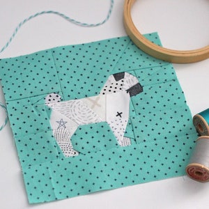 May include: A teal fabric square with a white and black paper pieced dog design. The fabric has a small white dot pattern. There are three spools of thread, two teal and one gray, on a white surface.