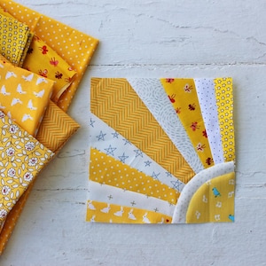 May include: A yellow and white fabric quilt block with a sunburst design. The block is made up of various yellow fabrics with different patterns, including polka dots, stars, and floral prints. The block is surrounded by a white border.