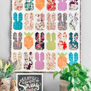 May include: A white quilt with a patchwork design of bunny rabbits and carrots. The quilt is hanging on a wall with a sign that says "Happy Spring" in front of it.