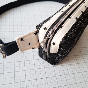 May include: A black and white fabric fanny pack with a zipper closure and a black adjustable strap. The fabric has a white background with black stars.