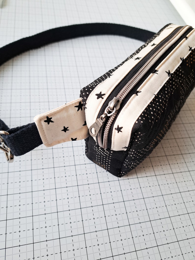 May include: A black and white fabric fanny pack with a zipper closure and a black adjustable strap. The fabric has a white background with black stars.