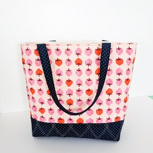 May include: A pink and white quilted tote bag with black straps and a black and white polka dot bottom. The bag is decorated with a pattern of red and pink strawberries.