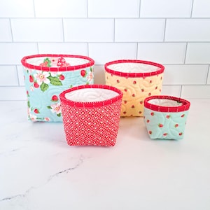 May include: A set of four fabric storage baskets in varying sizes. The baskets feature quilted textures and strawberry-themed patterns in light blue, yellow, and red, with white interiors and red trim along the top edges.