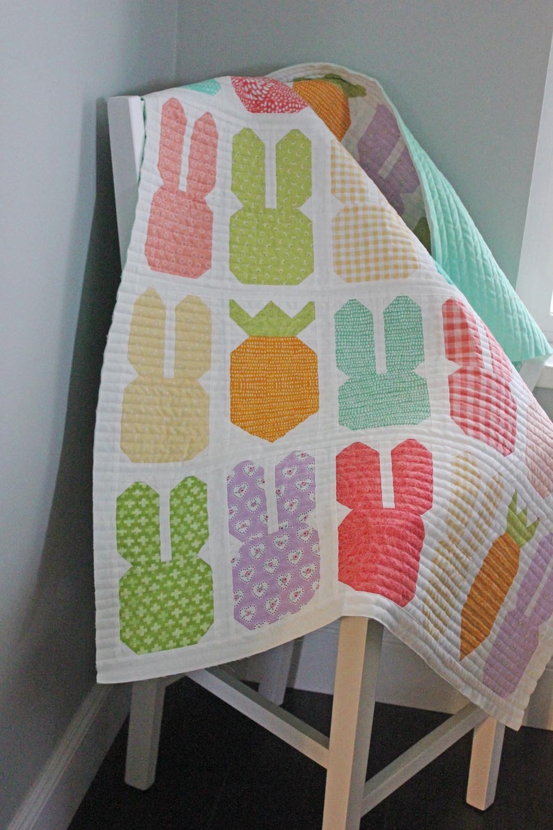 May include: A white quilt with a patchwork design featuring bunnies, carrots, and pineapples. The quilt is made with a variety of colorful fabrics, including pink, green, yellow, orange, and purple.