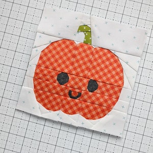 May include: A handmade fabric block with a smiling pumpkin design. The pumpkin is orange with a gingham pattern and black eyes. The stem is green with white polka dots. The background is white with small blue crosses.