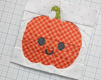 May include: A handmade fabric block with a smiling pumpkin design. The pumpkin is orange with a gingham pattern and black eyes. The stem is green with white polka dots. The background is white with small blue crosses.