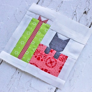 Peut inclure: Un bloc de courtepointe patchwork représentant un chat gris assis sur un cadeau rouge et blanc avec un cadeau vert et rouge à rayures sur le dessus.