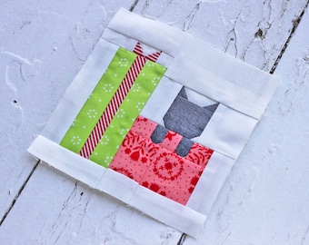 Op de afbeelding: Een patchwork quiltblok met een grijze kat die op een rood en wit cadeau zit met een groen en rood gestreept cadeau erbovenop.