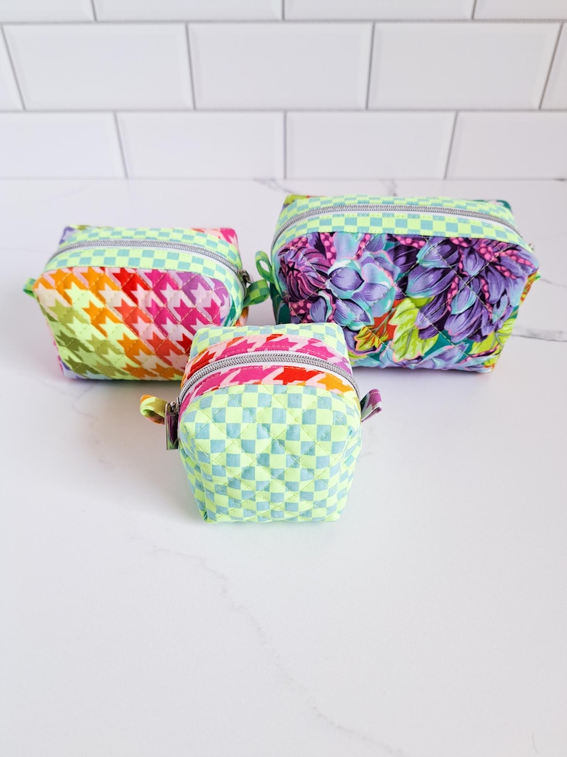 May include: Three quilted fabric zippered pouches in different patterns. The largest pouch is green with a floral print, the medium pouch is green and white checkered, and the smallest pouch is pink, orange, and yellow with a houndstooth pattern.