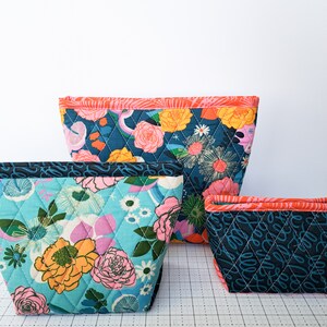May include: Three quilted fabric zippered pouches in different sizes and patterns. The largest pouch is teal blue with a floral print, the medium pouch is turquoise blue with a floral print, and the smallest pouch is black with a white and pink floral print. All pouches have a coral pink trim.
