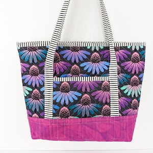 Op de afbeelding: Een zwarte quilted tote bag met een bloemenprint en een roze bodem. De tas heeft zwart-wit gestreepte hengsels.