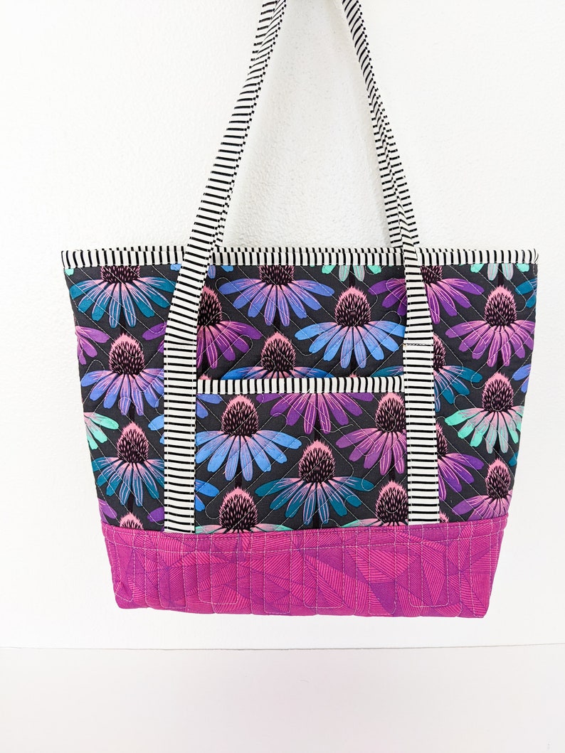 Op de afbeelding: Een zwarte quilted tote bag met een bloemenprint en een roze bodem. De tas heeft zwart-wit gestreepte hengsels.