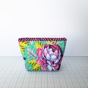 May include: A quilted makeup bag with a floral print featuring pink and yellow flowers on a teal background. The bag has a black and pink striped trim.