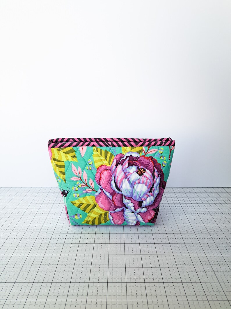 May include: A quilted makeup bag with a floral print featuring pink and yellow flowers on a teal background. The bag has a black and pink striped trim.