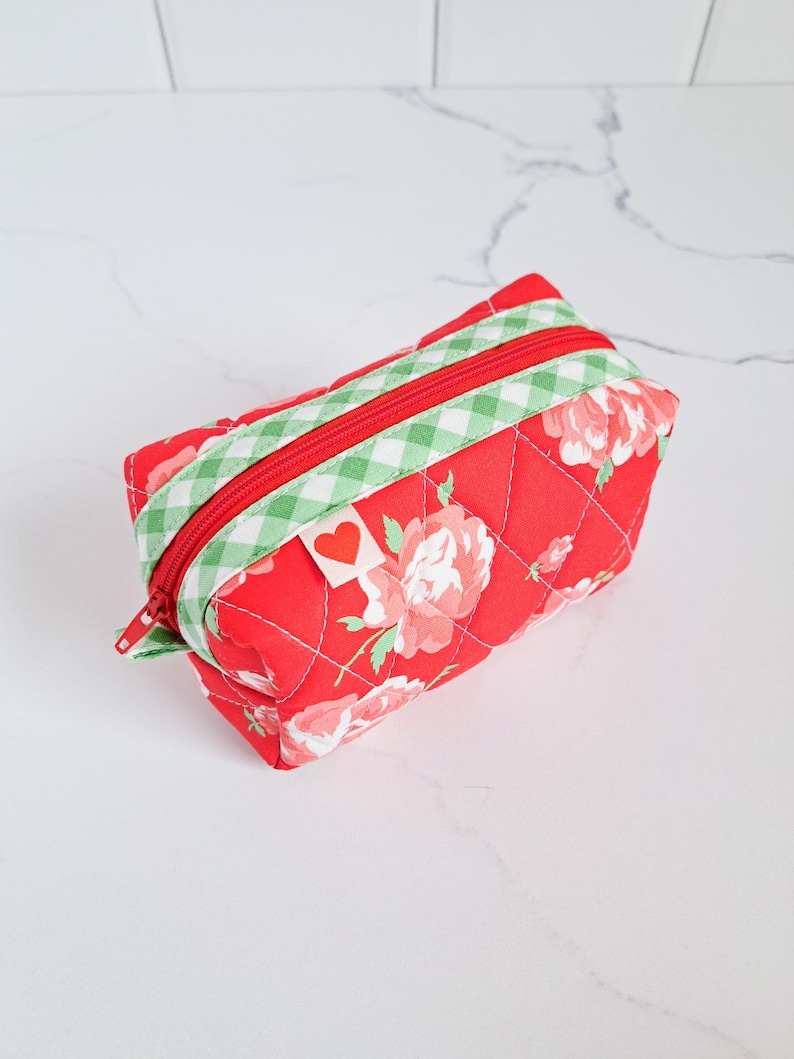 May include: Red quilted makeup bag with a green and white checkered trim and a red heart tag. The bag is decorated with a floral pattern.