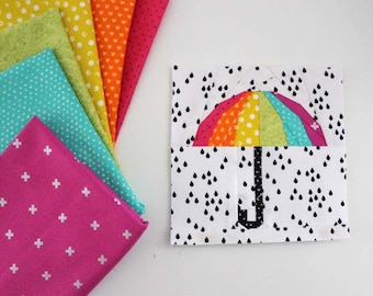 May include: A rainbow-colored fabric umbrella applique on a white background with black raindrops. The fabric pieces are arranged in a circle to form the umbrella shape. The umbrella is surrounded by a white border.