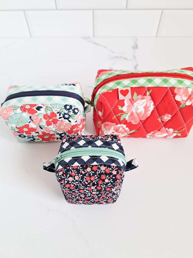 May include: Three quilted fabric zippered pouches in different sizes. The largest pouch is red with white and green floral print and a small red heart. The medium pouch is blue with a white and green floral print. The smallest pouch is black with a white and pink floral print.