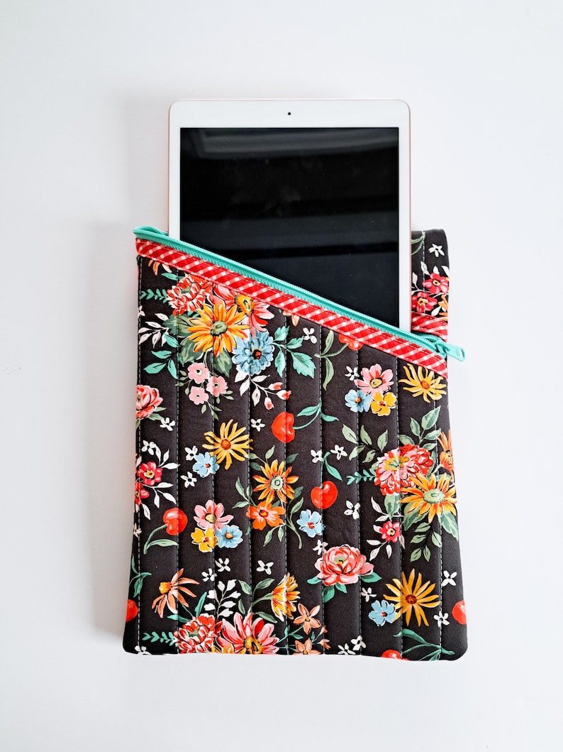 May include: A black quilted tablet case with a floral print and a red and white checkered zipper. The case is open and a tablet is partially visible inside.