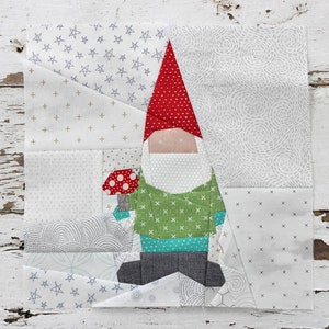 May include: A patchwork quilt block featuring a gnome holding a mushroom. The gnome is made of various fabrics in red, green, blue, and gray. The background is a white fabric with a variety of patterns.