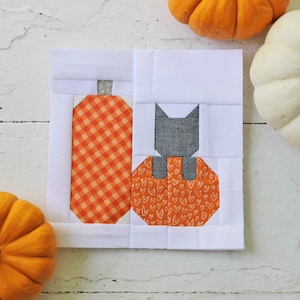 Może przedstawiać: Biały kwadratowy kawałek materiału z patchworkowym wzorem przedstawiającym czarnego kota siedzącego na dyni z pomarańczowymi i białymi sercami oraz dynią w pomarańczowo-białą kratkę. W tle znajdują się dwie dynie.