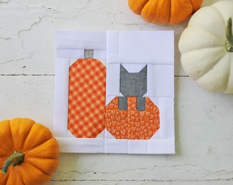May include: A white fabric square with a patchwork design featuring a black cat sitting on a pumpkin with orange and white hearts, and a pumpkin with orange and white checkered fabric. There are two pumpkins in the background.