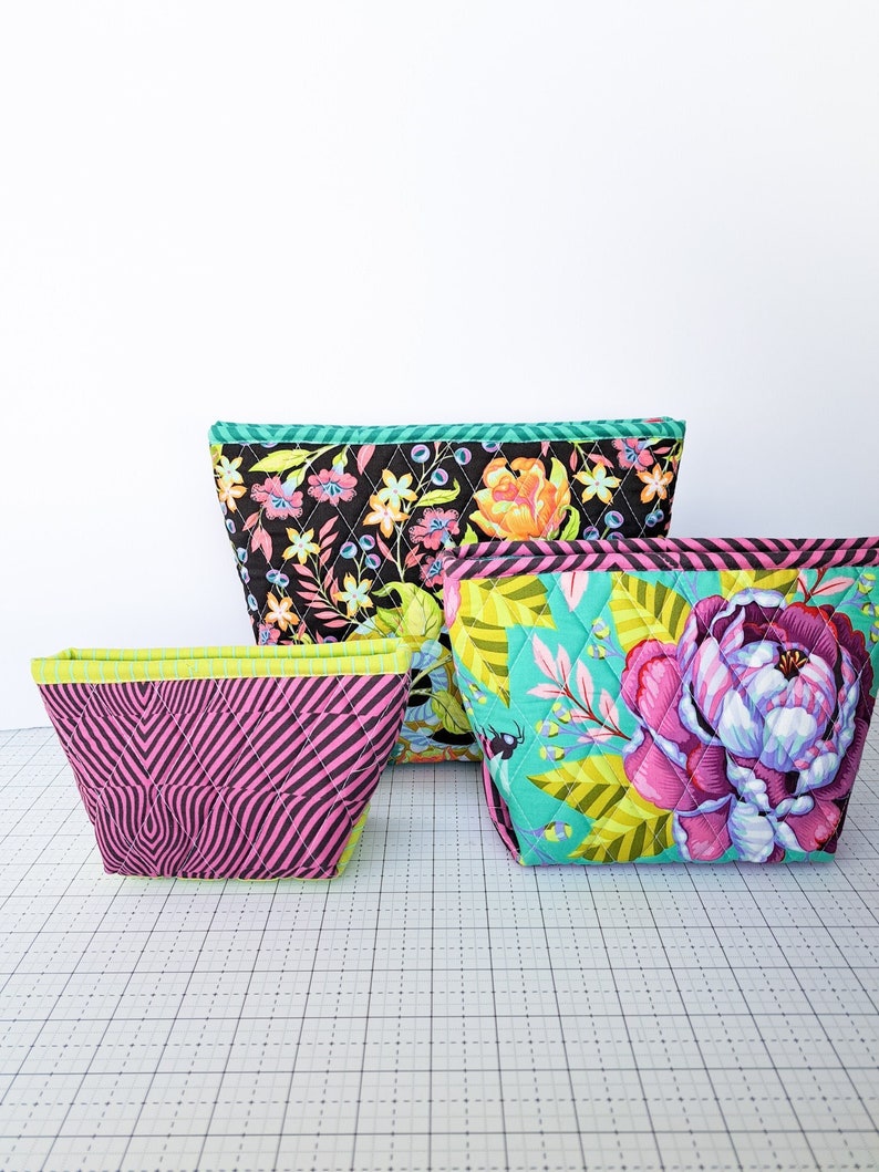May include: Three quilted fabric zippered pouches in different patterns and colors. The largest pouch is black with a floral print, the medium pouch is turquoise with a tropical leaf and flower print, and the smallest pouch is pink with a black and white geometric pattern.