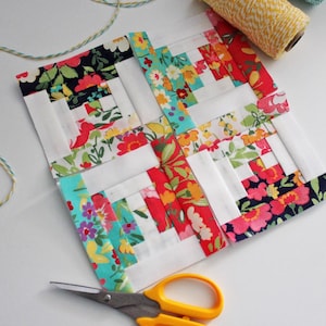 May include: A patchwork quilt block with a log cabin pattern. The block is made of white, red, pink, blue, and green fabric with floral prints. The block is surrounded by white fabric and is being trimmed with scissors.
