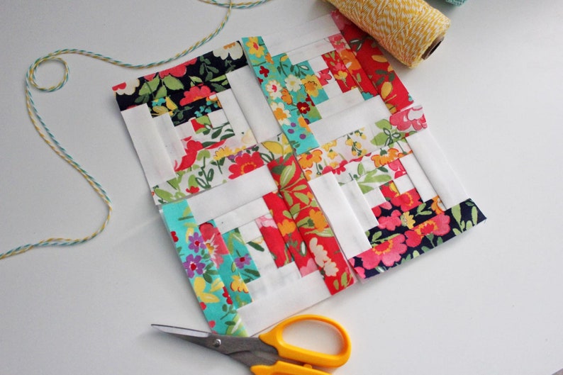 May include: A patchwork quilt block with a log cabin pattern. The block is made of white, red, pink, blue, and green fabric with floral prints. The block is surrounded by white fabric and is being trimmed with scissors.
