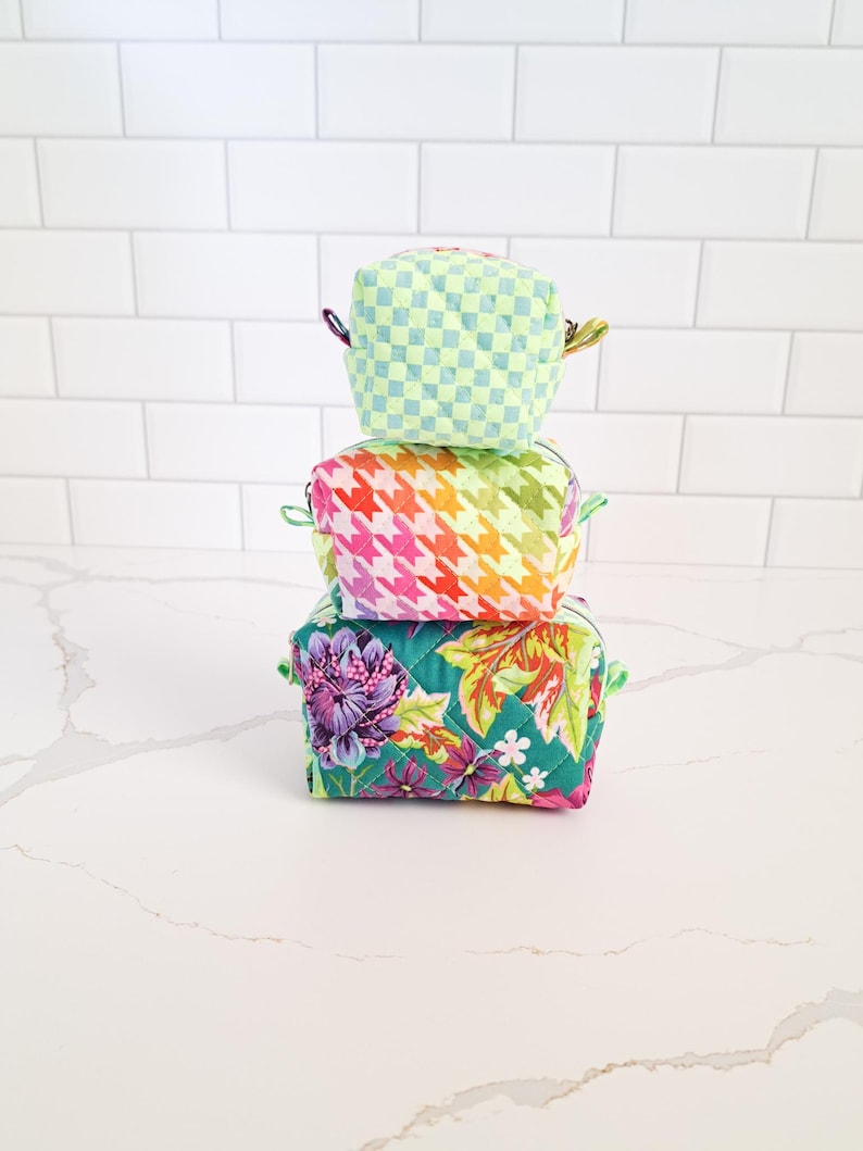 May include: Three quilted fabric pouches stacked on top of each other. The top pouch is light green with a white checkered pattern. The middle pouch is pink, orange, and white with a houndstooth pattern. The bottom pouch is green with a floral pattern.