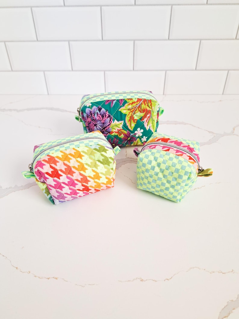 May include: Three quilted fabric pouches in different sizes and patterns. The largest pouch is green with a floral pattern, the medium pouch is rainbow with a houndstooth pattern, and the smallest pouch is light green with a checkered pattern.