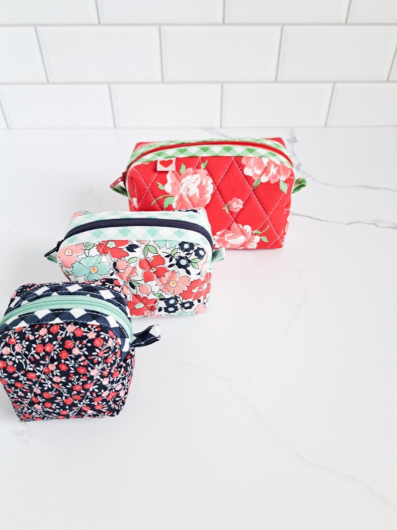 May include: Three quilted fabric zippered pouches in different sizes. The largest pouch is red with white and green gingham trim and a floral pattern. The medium pouch is blue and white with a floral pattern. The smallest pouch is black and red with a floral pattern.