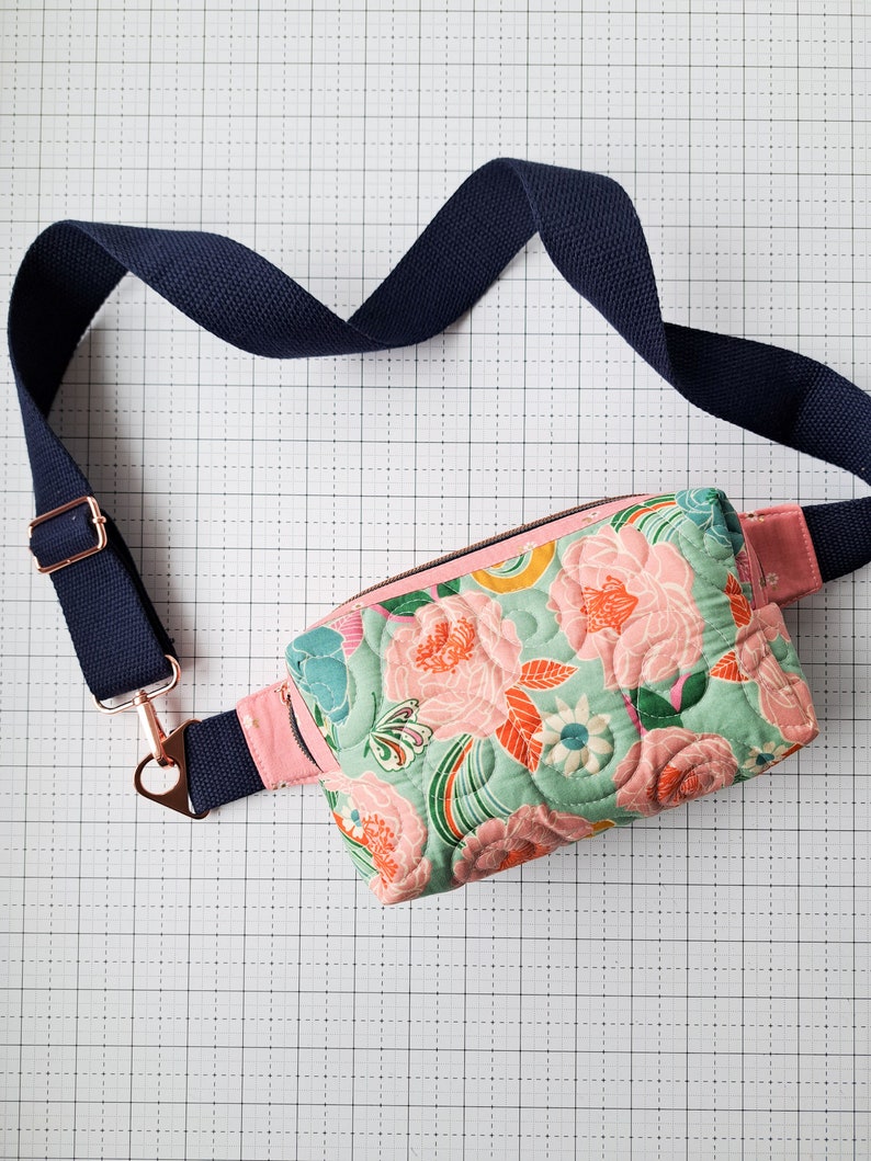 May include: A quilted floral fanny pack with a pink and green floral print. The pack has a navy blue adjustable strap with a silver buckle and a gold-toned zipper.