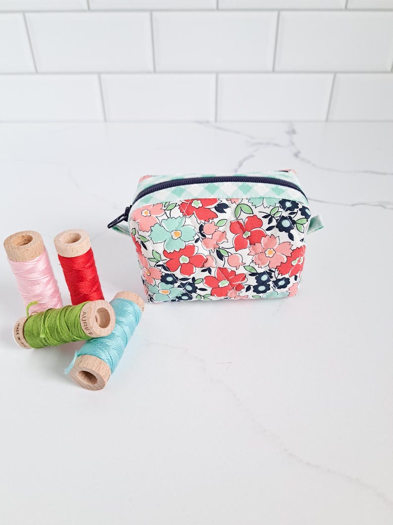 May include: A small floral print zippered pouch with a blue and white checkered trim. The pouch is sitting on a white marble surface with four spools of thread in various colors.