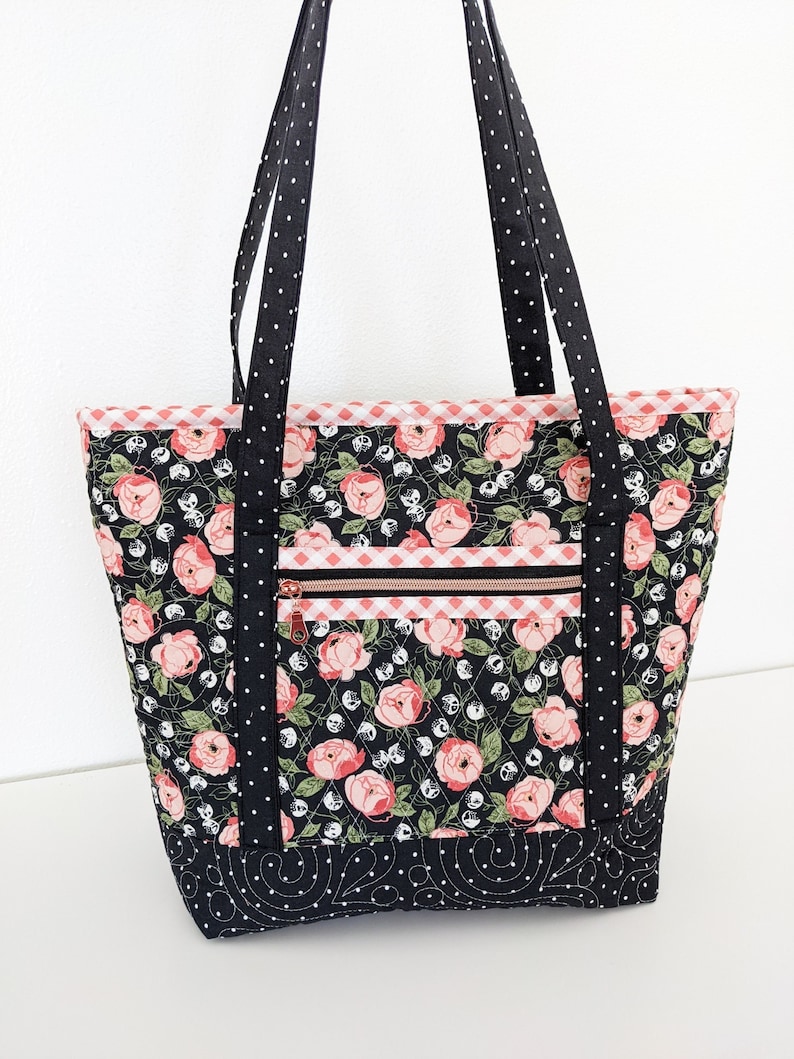 Op de afbeelding: Zwarte tote bag met een bloemenprint met roze rozen en witte stippen. De tas heeft een ritsvak en zwart-witte gestippelde banden.
