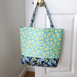 May include: A blue and green floral tote bag with yellow flowers and a navy blue trim. The bag has two straps and is hanging on a white door.